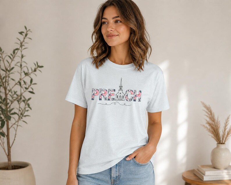 以下が含まれることがあります： 胸元に「PREACH」の文字がプリントされたライトブルーの半袖Tシャツ。文字はダークな花柄で埋め尽くされており、「A」の文字は教会の尖塔の線画に置き換えられ、その下に装飾的なラインが引かれています。