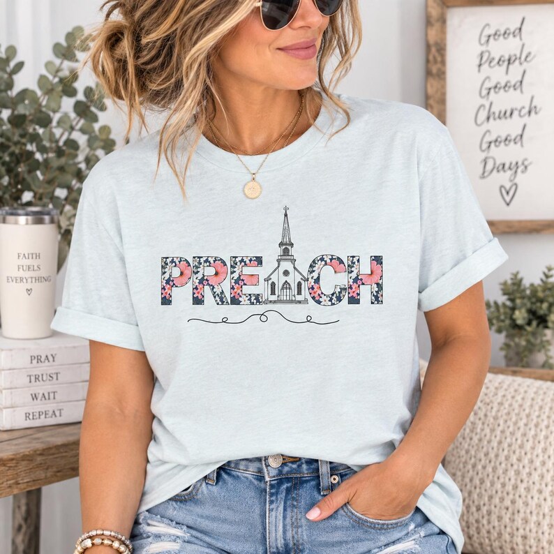 以下が含まれることがあります： 花柄の太いブロック体で「PREACH」と書かれたライトブルーのTシャツ。Aの文字は教会の線画に置き換えられています。テキストの下には、細い装飾的な水平線が配置されています。