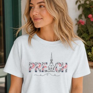 以下が含まれることがあります： ピンクのアクセントが入ったダークな花柄で「PREACH」という文字がプリントされた白い半袖Tシャツ。文字の「A」は教会の尖塔の線画イラストに置き換えられています。テキストの下には装飾的な渦巻き線が描かれています。
