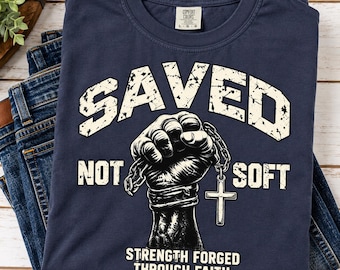 以下が含まれることがあります： ネイビーブルーのTシャツで、十字架のペンダントが付いた鎖を握りしめた拳のグラフィックが描かれています。SAVED NOT SOFTおよびSTRENGTH FORGED THROUGH FAITHという文字が、かすれた白のフォントでプリントされています。Comfort Colorsのブランドラベルが付いています。