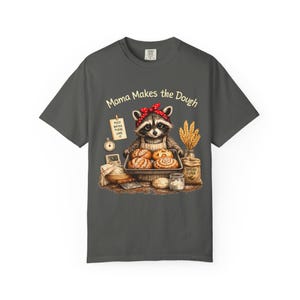 Peut inclure: Un t-shirt gris anthracite avec une illustration de raton laveur portant un bandana rouge et un tablier, tenant un plateau de quatre pains. Le texte indique "Mama Makes the Dough" avec une note "FEED WATER FLOUR LOVE" et un sac de farine.