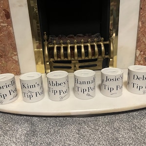 Könnte beinhalten: Sechs weiße Trinkgeld-Töpfe mit Namen und der Aufschrift "Tip Pot". Jeder Topf hat einen Schlitz und eine kleine Grafik eines Salonwerkzeugs. Die Töpfe sind auf einer weißen Oberfläche angeordnet.