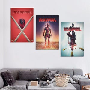 May include: Three Deadpool film posters on a white wall. The posters show the Deadpool character in action poses, with the film title in red. The posters are framed and hung above a grey sofa in a living room setting.
