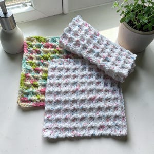 May include: Three handmade crochet washcloths. One is white with flecks of pink, blue, and yellow. Another is green, pink, and yellow. The third is white with flecks of pink and blue. The washcloths are on a windowsill.