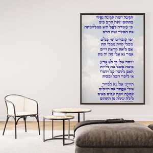 May include: A framed print with Hebrew text in blue against a cloudy sky background. The print hangs on a white wall above a gray ottoman. A white chair and two small round tables are in front of the wall. A potted plant is on the left.