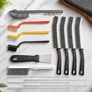 May include: A collection of cleaning tools, including brushes with red, yellow, and black handles, a black scraper, and several long, thin brushes. The tools are arranged on a white marble surface.