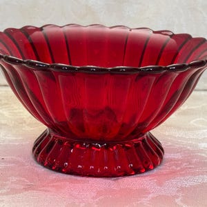 May include: A deep red glass bowl featuring a scalloped rim and vertical ribbed detailing along the sides. The bowl sits on a circular pedestal base that mirrors the ribbed pattern found on the main body of the vessel.