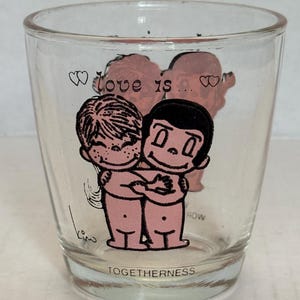 May include: Clear glass shot glass with a cartoon illustration of two figures embracing. The words "love is..." and "TOGETHERNESS" are printed on the glass. The figures are pink with black outlines.