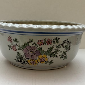 May include: A white ceramic bowl with a scalloped rim and a blue horizontal line near the top. The exterior features a floral pattern with purple, yellow, and pink flowers, green leaves, and dark green stems painted on the side of the vessel.