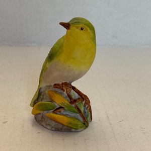 May include: A small, ceramic bird figurine. The bird has yellow and green plumage, a brown beak, and is perched on a gray rock with yellow and green leaves. The figurine is a decorative item.