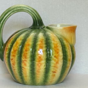 May include: A ceramic pitcher shaped like a pumpkin with a glossy finish. The body features vertical ridges in shades of green and yellow. A curved green handle extends from the top, and a wide, yellow-toned spout is positioned on the side.