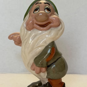 May include: A ceramic figurine of a character from a popular animated film. The figure is depicted with a white beard, a green hat, and a green and brown outfit. The figurine has a smiling expression.