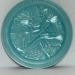 May include: A round, turquoise ceramic plate with a raised relief design. The plate features a seated figure working at a table, with architectural elements in the background. The rim has a decorative, repeating pattern.