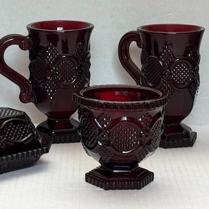 May include: A collection of deep red pressed glass tableware featuring a butter dish with a lid, two handled mugs, four stemmed goblets, and one small bowl. Each piece displays a consistent geometric pattern with circular and diamond motifs.