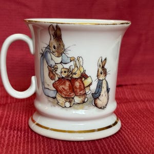 May include: A white ceramic mug featuring a colorful illustration of several rabbits, including one wearing a blue coat and another in a red jacket. The mug has a curved handle and a gold-colored trim along the top rim and the base of the vessel.