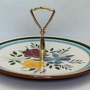 May include: A round ceramic serving tray with a cream-colored base featuring a floral pattern of yellow, purple, and blue flowers with green leaves. The rim has a blue braided border and a dark green edge. A gold-tone metal handle is centered.