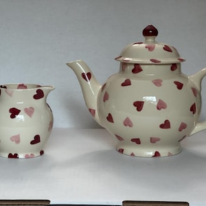May include: A cream-colored ceramic teapot and matching creamer pitcher, both decorated with a repeating pattern of small, hand-painted hearts in varying shades of dark red, maroon, and light pink. The teapot features a lid with a knob.