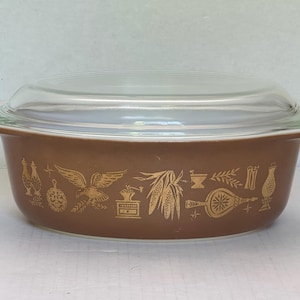 May include: A brown oval casserole dish with a clear glass lid. The exterior features a gold-colored pattern depicting an eagle, corn, a coffee grinder, a mortar and pestle, and other kitchen-related motifs printed around the center.