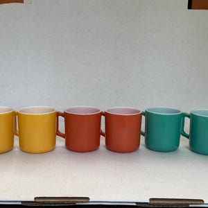 May include: A set of six ceramic mugs arranged in a row, featuring two yellow mugs, two orange mugs, and two teal mugs. Each mug has a smooth, solid-colored exterior, a rounded handle on the side, and a white interior rim visible at the top.