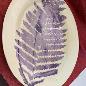 May include: An oval-shaped ceramic platter with a cream-colored base and a large, detailed purple fern leaf print centered on the surface. A small portion of a green measuring tape is visible at the bottom left corner, showing metric units.