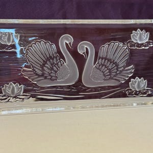 May include: A rectangular glass serving tray with etched swan and water lily designs. The tray features two swans facing each other, surrounded by water lilies. The tray has handles on each side and is clear with a dark background.