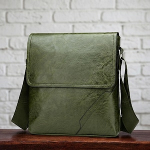 May include: Olive green leather messenger bag with a flap closure and adjustable shoulder strap. The bag has a textured finish and a rectangular shape. The strap is attached with silver-colored hardware. The bag is sitting on a wooden surface.