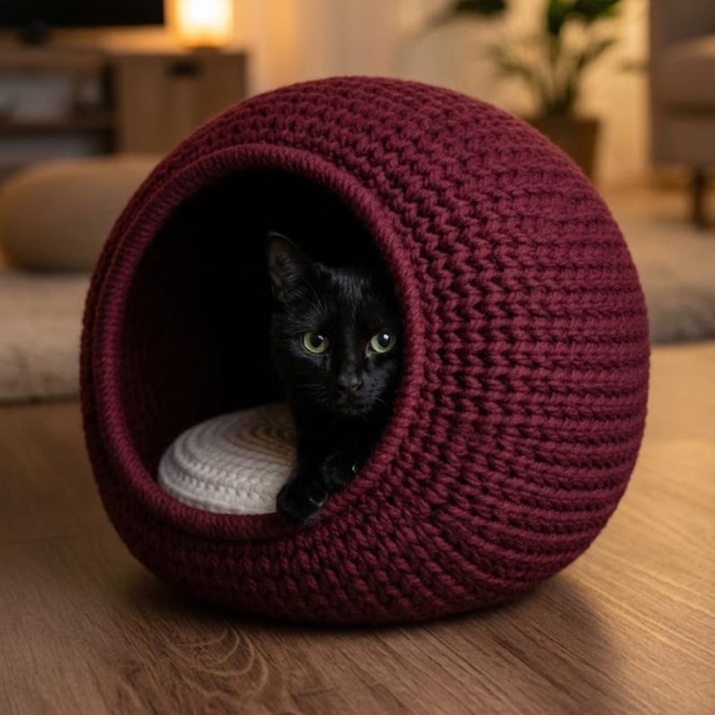 May include: A burgundy-colored, spherical cat bed made of thick, woven material. A black cat with green eyes is nestled inside, resting on a cream-colored cushion. The bed is placed on a wooden floor, creating a cozy and inviting space for the cat.