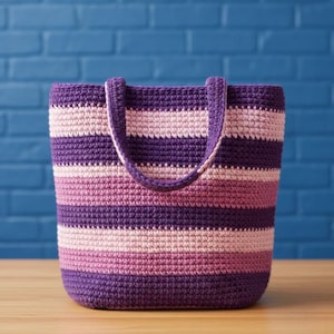 May include: A handmade crochet tote bag with horizontal stripes in shades of purple and pink. The bag has a rectangular shape with a sturdy handle. The bag is sitting on a wooden surface with a blue brick wall in the background.