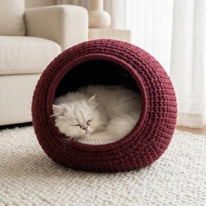 May include: A sleeping white cat inside a burgundy crochet pet bed. The round bed has a dark interior and sits on a white rug. The cat is curled up, with its eyes closed.