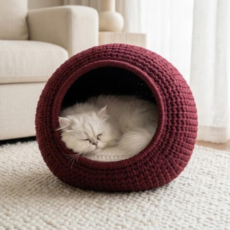 May include: A sleeping white cat inside a burgundy crochet pet bed. The round bed has a dark interior and sits on a white rug. The cat is curled up, with its eyes closed.