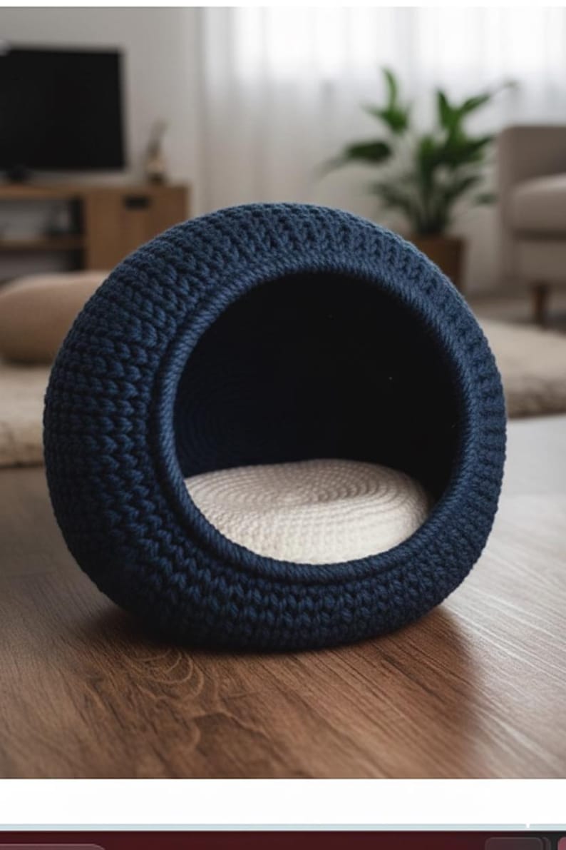 May include: A navy blue, round pet bed with a woven texture. The bed has a circular opening and a cream-colored, woven interior cushion. The bed is placed on a wooden floor, suggesting a cozy indoor setting for pets.