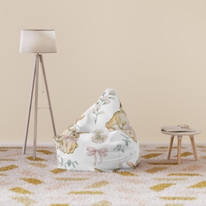 May include: A white beanbag chair with a pattern of watercolor bunnies, pink bows, and floral accents. The chair is in a room with a floor lamp, a small side table, and a patterned rug.