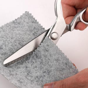 May include: Silver dressmaking shears cutting a piece of grey felt. The shears have a serrated blade and large finger holes. The felt is being cut with a zig-zag pattern. The felt is grey.
