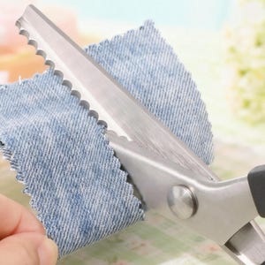 May include: A close-up shot of silver-coloured, serrated scissors cutting a piece of blue denim fabric. The scissors have black handles. The fabric is being cut with a zig-zag pattern. The scissors are held by a person's hand.
