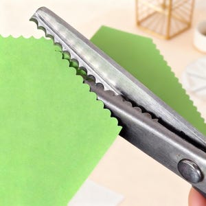 Pode incluir: Close-up de tesouras prateadas e serrilhadas cortando um pedaço de papel verde brilhante. As tesouras têm um design de lâmina ondulada, criando uma borda decorativa. Papel verde adicional é visível no fundo.