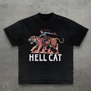Könnte beinhalten: Schwarzes T-Shirt mit einer Grafik einer Frau, die auf einem Tiger über Flammen reitet, und dem weißen Text "HELL CAT". Das Design hat einen Vintage-Look. Ein einzigartiges Grafik-T-Shirt.