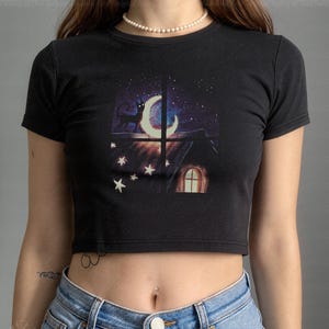 Op de afbeelding: Zwarte crop top met een illustratie van een nachtscène. Het ontwerp toont een zwarte kat, een maan en sterren door een raam, met een huis en een verlicht raam. De top wordt gedragen met lichtblauwe denim jeans. Een casual kledingstuk.