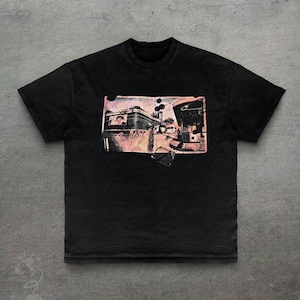 Pode incluir: Uma t-shirt preta de manga curta com uma impressão gráfica retangular no peito. A impressão exibe uma cena de estilo vintage de um restaurante ou edifício com uma paleta de cores rosa e castanho desbotadas, incluindo um pequeno retrato.