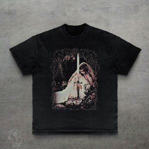 May include: Black t-shirt featuring a detailed graphic of a figure holding a sword, set within an ornate frame. The design has a vintage, distressed look. The shirt's fabric appears soft, and the overall aesthetic is gothic and artistic.