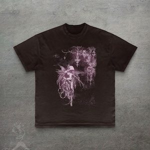 May include: Dark brown t-shirt with a purple fairy illustration. The design features a winged figure with a flowing gown and gothic details. The tee has a crew neck and short sleeves. The image has a vintage look.