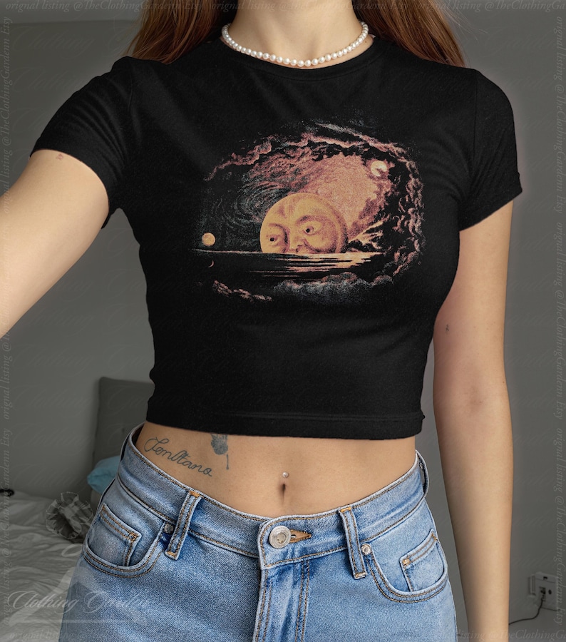 May include: Black crop top featuring a surreal illustration of a moon with a face emerging from water, surrounded by clouds. The artwork is in shades of yellow, pink, and brown. The shirt has short sleeves and a crew neck.