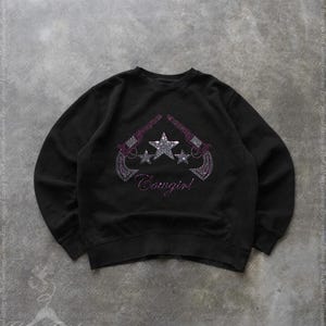 Könnte beinhalten: Schwarzes Sweatshirt mit einem Strass-Design, das zwei Pistolen, Sterne und das Wort "Cowgirl" in Rosa zeigt. Das Design befindet sich mittig auf der Vorderseite des Rundhals-Sweatshirts. Das Sweatshirt ist aus weichem Material.