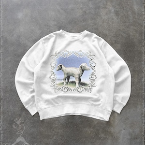 May include: White sweatshirt featuring a two-headed lamb illustration within a decorative frame. The artwork depicts the lamb standing on grass with a blue sky background. The sweatshirt has a crew neck and long sleeves.