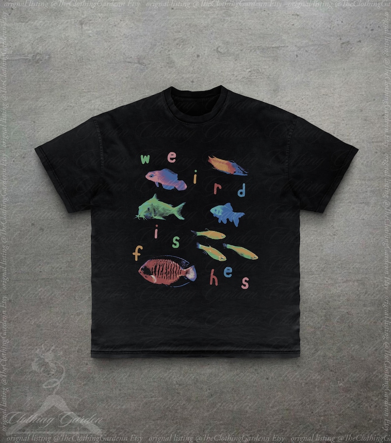 Puede incluir: Camiseta negra con ilustraciones coloridas de varias especies de peces y el texto "weird fishes" en una fuente juguetona. El dise&ntilde;o incluye peces en tonos azules, verdes, naranjas y rojos, creando un gr&aacute;fico vibrante y llamativo.