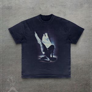 Pode incluir: Camiseta azul marinho escuro com um gr&aacute;fico desgastado de um anjo ajoelhado com grandes asas. O anjo est&aacute; em uma luz brilhante e brilhante. A camiseta tem gola redonda cl&aacute;ssica e mangas curtas.