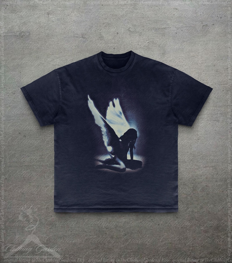 Pode incluir: Camiseta azul marinho escuro com um gr&aacute;fico desgastado de um anjo ajoelhado com grandes asas. O anjo est&aacute; em uma luz brilhante e brilhante. A camiseta tem gola redonda cl&aacute;ssica e mangas curtas.