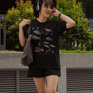 Puede incluir: Camiseta negra con las palabras "weird fishes" e ilustraciones de peces de colores. La camiseta la lleva una persona con auriculares y una bolsa de mano gris.