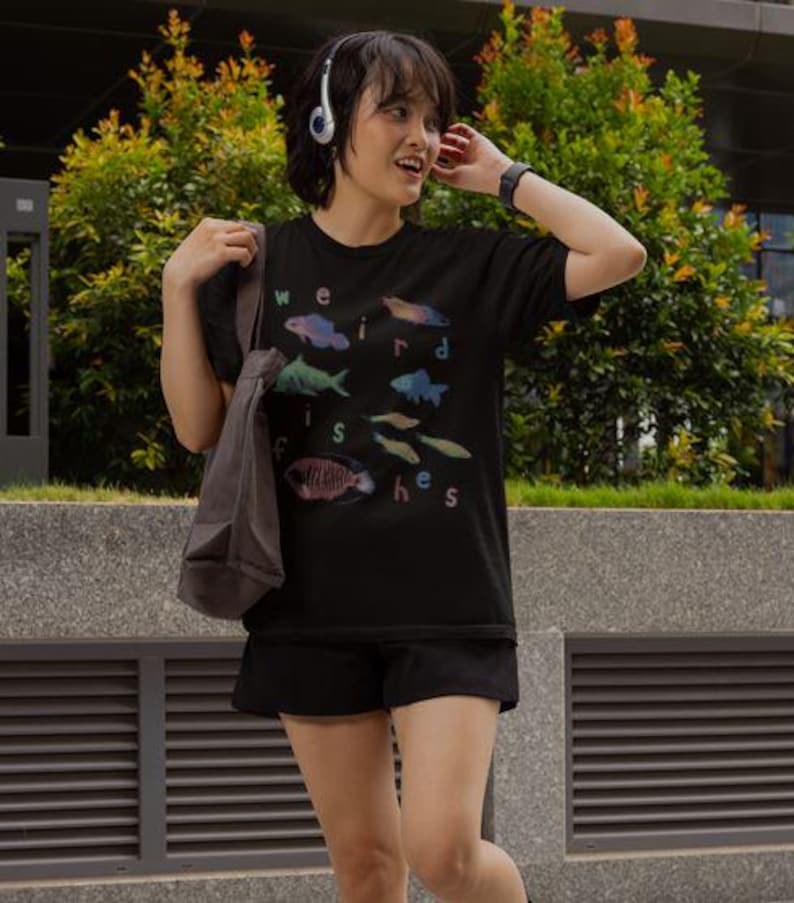 Puede incluir: Camiseta negra con las palabras "weird fishes" e ilustraciones de peces de colores. La camiseta la lleva una persona con auriculares y una bolsa de mano gris.