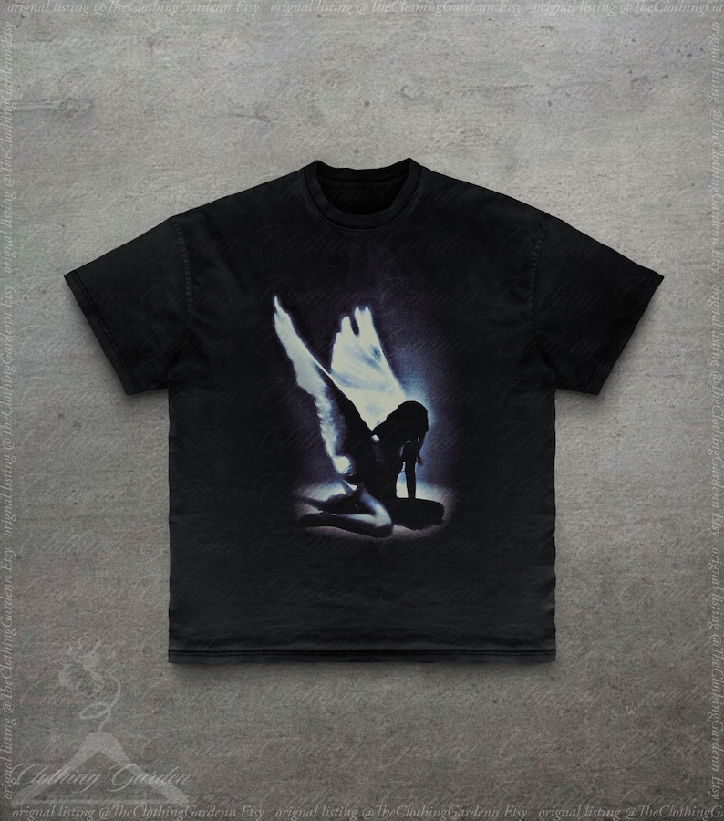 Pode incluir: Camiseta preta com um gr&aacute;fico desgastado de um anjo ajoelhado com grandes asas. O anjo &eacute; retratado em tons de azul e branco contra um fundo escuro. A camiseta tem gola redonda e mangas curtas.