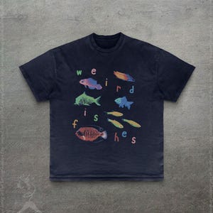 Puede incluir: Camiseta azul marino con ilustraciones coloridas de varios peces y el texto "weird fishes" en una fuente l&uacute;dica. El dise&ntilde;o incluye peces en tonos azules, naranjas, verdes y amarillos.